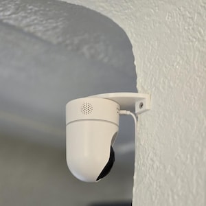 Peut inclure: Une caméra de sécurité blanche montée sur un mur blanc. La caméra a un corps arrondi avec un objectif noir et un petit haut-parleur. Un câble blanc est connecté à la caméra.