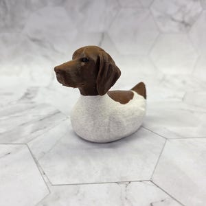 Peut inclure: Une petite figurine en forme de canard en caoutchouc, mais dotée de la tête et des oreilles d'un chien marron. Le corps de la figurine est blanc avec une surface texturée, tandis que la tête est d'une couleur marron unie avec des yeux sombres.
