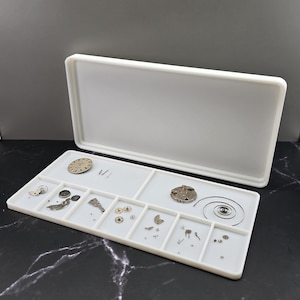 Può includere: Un vassoio rettangolare bianco per parti di riparazione di orologi con coperchio, contenente vari componenti di orologi. Il vassoio è diviso in scomparti che contengono ingranaggi, quadranti e altre piccole parti. Lo sfondo è scuro.