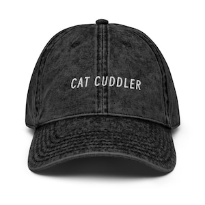 May include: A black acid-wash baseball cap featuring the embroidered white text CAT CUDDLER centered on the front panel. The hat has a structured crown, a curved brim, and a button at the top, showcasing a distressed fabric texture throughout.