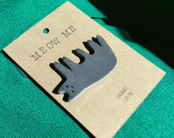 May include: A handmade gray clay pin in the shape of a cat, attached to a brown tag. The tag reads "MEOW ME" and "HANDMADE CLAY PIN". The pin is on a green surface.