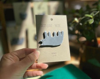 May include: A gray cat-shaped clay pin on a brown card with the text "MEOW ME". The pin is handmade and has a simple, stylized design. The card also has the text "HANDMADE CLAY PIN".