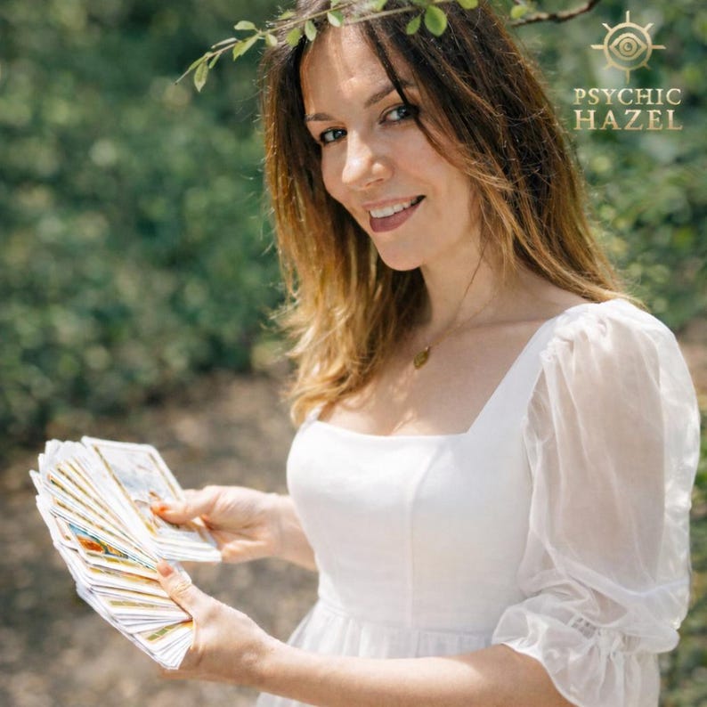 May include: A woman holding a deck of tarot cards, smiling at the camera. She wears a white dress with sheer sleeves and a leafy headpiece. The text "PSYCHIC HAZEL" is visible in the upper right corner.