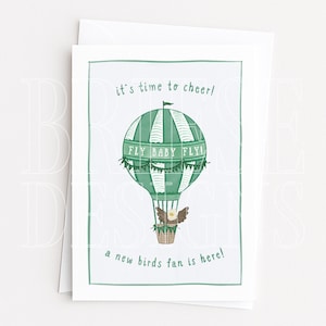 May include: A greeting card featuring a green and white striped hot air balloon with the text "it's time to cheer!", "FLY BABY FLY!", and "a new birds fan is here!". A small eagle with spread wings sits in the brown basket beneath the balloon.