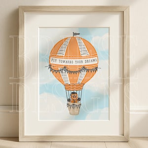 May include: Framed art print featuring an orange hot air balloon with gray stripes and the text "FLY TOWARDS YOUR DREAMS." A cartoon character is in the basket. The background is a light blue sky with clouds. The print is in a light wood frame.