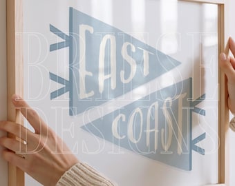 May include: A framed print featuring two light blue pennant flags with the words "EAST COAST" in white. The artwork is in a light wooden frame. The image is held by a person.