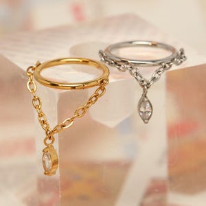 May include: Two rings with chain detailing and teardrop-shaped gems. One ring is gold-coloured with a matching chain and gem, the other is silver-coloured. The rings are displayed on a clear, square block.
