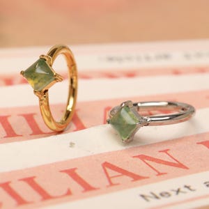 Pode incluir: Dois anéis com pedras verdes de corte quadrado. Um anel é dourado, o outro prateado. Os anéis estão sobre uma superfície listrada rosa e branca com texto. Os anéis são exibidos em close-up.