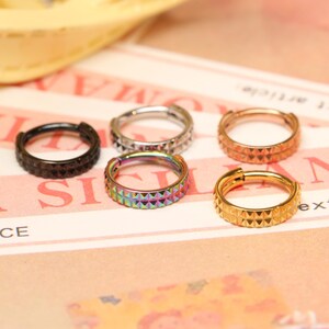 Puede incluir: Cinco pendientes de aro de diferentes colores con un diseño texturizado. Los aros son de metal y tienen un acabado brillante. Los colores son negro, plata, oro rosa, arcoíris y oro.