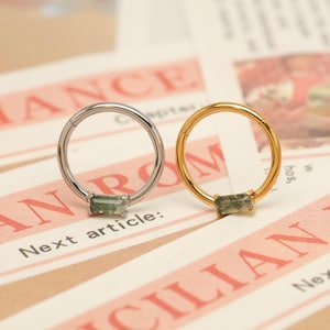 May include: Two hoop earrings, one silver and one gold, each featuring a rectangular green gemstone. The earrings are displayed on a newspaper-like background with the text "ANCE" and "ROM". The text "Next article:" is also visible.