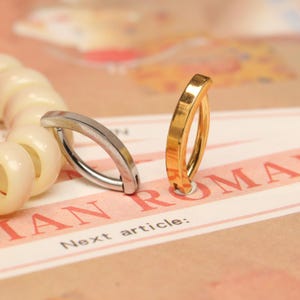 Puede incluir: Dos piercings para el ombligo curvados, uno plateado y otro dorado, se muestran sobre una superficie impresa. Los anillos tienen un diseño simple y elegante. El texto "IAN ROMA" y "Next article:" es visible en la superficie.