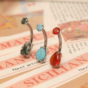 May include: Three belly button rings with curved silver-tone bars. Each ring features a different gemstone: green, turquoise, and red-orange. The gemstones are teardrop-shaped and set in silver-tone metal.