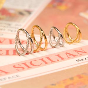 May include: Four silver and gold hoop earrings with a twisted rope design. The earrings are arranged in a row on a white and red surface.