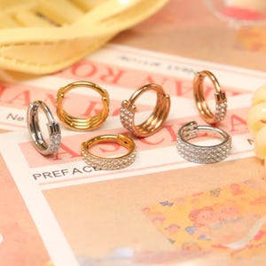 Puede incluir: Cinco pendientes de aro con una fila de piedras de circonita cúbica transparentes. Los aros son de plata, oro y oro rosa.