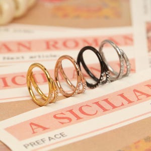 May include: Four rings in gold, rose gold, black, and silver, each with a double band design. The rings are adorned with small, sparkling stones. The rings are displayed on a printed paper background.