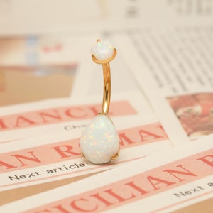 May include: A gold-coloured belly button ring featuring two white opal stones. The top stone is round, and the bottom stone is teardrop-shaped. The curved bar connects the two stones. The jewellery is set against a newspaper background.