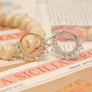May include: Two decorative nose rings. One is gold-toned, the other is silver-toned. Both are circular with a sunburst design of clear, square-cut gemstones. The rings are displayed on a printed background with the words "A SICILIA" visible.
