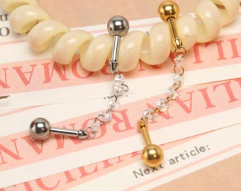 Op de afbeelding: Twee barbell oorbellen, één zilver en één goud, elk met een bal aan één uiteinde en een decoratieve ketting van heldere kralen. De oorbellen worden gepresenteerd op een roze en wit gestreept oppervlak met tekst.