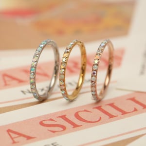 Puede incluir: Tres anillos delicados, cada uno adornado con una fila de pequeñas piedras preciosas iridiscentes. Un anillo es plateado, otro dorado y el tercero es de oro rosa. Los anillos se exhiben sobre una superficie rosa y blanca.