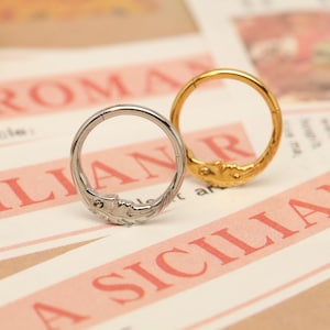 May include: Two decorative rings, one silver and one gold, are displayed. The silver ring features a detailed leaf design, while the gold ring has an ornate, textured pattern. Both rings are circular and appear to be made of metal.