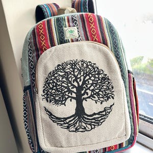 Puede incluir: Mochila colorida con diseño de árbol de la vida. El bolsillo delantero es de color crema con un diseño de árbol negro. El cuerpo principal tiene un patrón de rayas en rojo, verde y azul. Las correas y el asa superior también son rayadas.