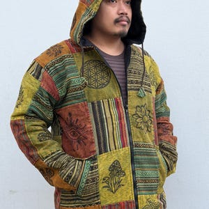 May include: A patchwork hooded jacket with a full-length zip. The jacket features a variety of colourful fabric squares with printed designs, including an eye, flower, and geometric patterns. The jacket has a black lining and a drawstring hood.