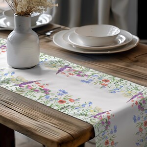 Puede incluir: Un camino de mesa blanco con un patrón floral que presenta una variedad de flores silvestres coloridas. El camino de mesa está colocado sobre una mesa de madera con un jarrón de flores secas y un tazón blanco.