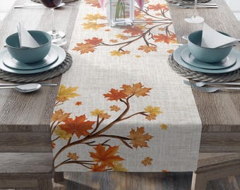 May include: A beige table runner with a repeating pattern of autumn leaves in shades of orange and yellow. The leaves are arranged on brown branches. The runner is set on a wooden table, with place settings and a vase of pink flowers.