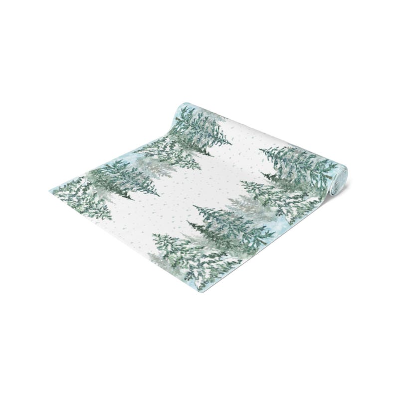 Puede incluir: Un camino de mesa blanco enrollado con un dise&ntilde;o de acuarela de &aacute;rboles de hoja perenne y nieve que cae. Los &aacute;rboles son en tonos verdes y azules, creando una escena de bosque invernal. El camino de mesa est&aacute; parcialmente enrollado.