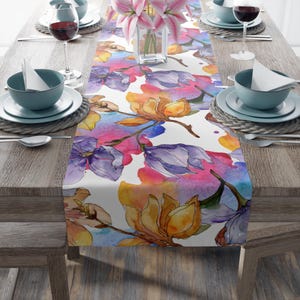 Puede incluir: Un camino de mesa blanco con un diseño floral de acuarela con flores moradas, amarillas y rosas. El camino de mesa está sobre una mesa de madera con dos cubiertos a cada lado.