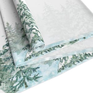 Puede incluir: Una tela enrollada con una escena de bosque invernal. El dise&ntilde;o presenta &aacute;rboles de hoja perenne cubiertos de nieve en tonos verdes y blancos, con un borde azul claro. La tela est&aacute; parcialmente desenrollada, revelando la impresi&oacute;n detallada.