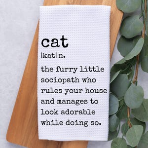 May include: White kitchen towel with black text that reads "cat |kat| n. the furry little sociopath who rules your house and manages to look adorable while doing so."