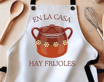 May include: White kitchen apron with black straps and a brown pot with yellow flowers. The text on the apron reads "En la casa hay frijoles".