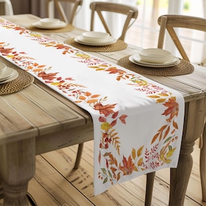 May include: White table runner with a fall leaf design. The runner features orange, red, and yellow leaves with green stems and berries. The runner is on a wooden table with two place settings.