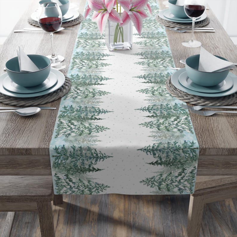 Puede incluir: Un camino de mesa blanco con un dise&ntilde;o de acuarela de pinos verdes en los bordes. El camino de mesa est&aacute; sobre una mesa de madera con cuencos, platos y copas de vino de color turquesa. Los lirios rosas est&aacute;n en un jarr&oacute;n transparente.