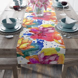 Puede incluir: Un camino de mesa colorido con un diseño de acuarela de flores rosas, amarillas y naranjas con hojas verdes y salpicaduras de pintura azul y amarilla sobre un fondo blanco.