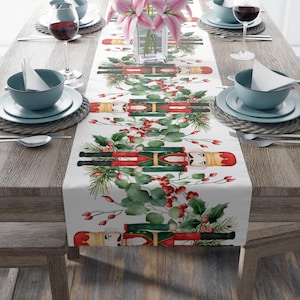 May include: A white table runner featuring a repeating pattern of toy nutcrackers in red, green, and gold. The design includes sprigs of holly and red berries, creating a festive holiday theme. The runner is placed on a wooden table.
