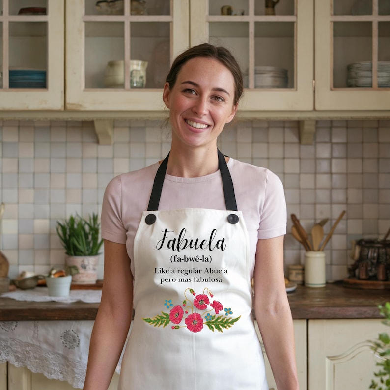 May include: White apron with black straps and a floral design. The apron has the text "Fabuela (fa-bw&ecirc;-la) Like a regular Abuela pero mas fabulosa" printed on it.