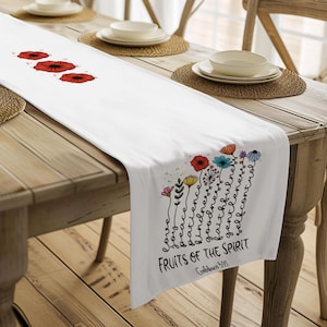 May include: White table runner with red poppy flowers and a colorful floral design. The runner features the text "Fruits of the Spirit" and is displayed on a wooden table with place settings.