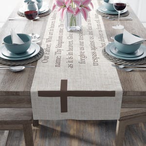 May include: A beige linen table runner with a brown cross design and the Lord's Prayer printed in brown text. The runner is on a wooden table with a vase of pink flowers and place settings.