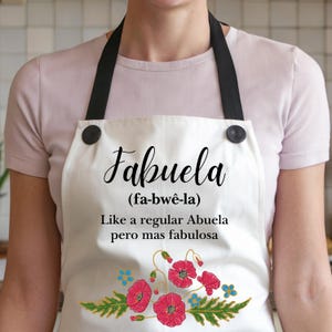 May include: White apron with black straps and buttons. The apron has a floral design with red poppies and green leaves. The text on the apron reads "Fabuela (fa-bw&ecirc;-la) Like a regular Abuela pero mas fabulosa".