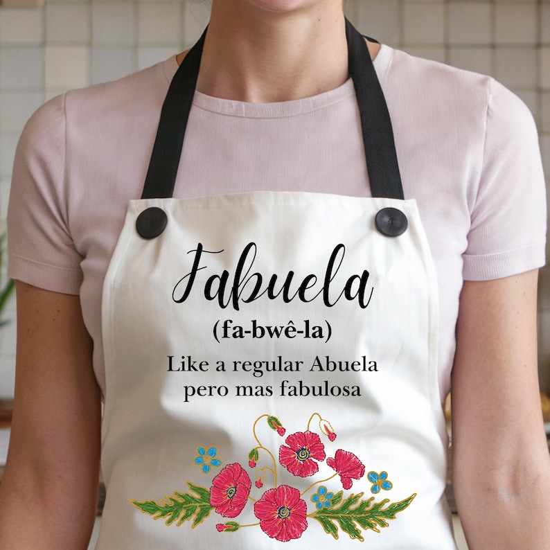 May include: White apron with black straps and buttons. The apron has a floral design with red poppies and green leaves. The text on the apron reads "Fabuela (fa-bw&ecirc;-la) Like a regular Abuela pero mas fabulosa".
