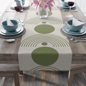 May include: A beige table runner with a geometric pattern of green circles and arches. The runner is on a wooden table with place settings on either side.