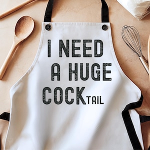 May include: White apron with black straps and the text "I NEED A HUGE COCKTAIL" in a bold, distressed font. The apron is on a light brown surface, with a wooden spoon and whisk visible.