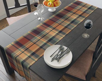 May include: A plaid table runner in shades of orange, red, green, and tan, displayed on a dark wooden table. The table is set with white plates, silverware, wine glasses, and a bowl of apples, creating a dining setting.