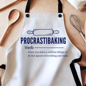 May include: White apron with black straps featuring the text "PROCRASTIBAKING" in blue, along with a rolling pin illustration. The text below defines the word as a verb, describing the act of baking instead of doing other tasks.