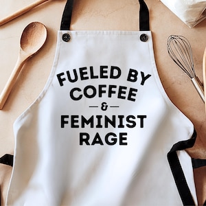 May include: White apron with black straps and the text "Fueled by Coffee & Feminist Rage." The apron has black accents and is likely made of a durable fabric. A wooden spoon and whisk are in the background.