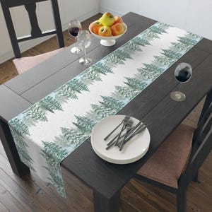 Puede incluir: Un camino de mesa blanco con un patr&oacute;n repetido de pinos verdes y azules en acuarela. El camino de mesa est&aacute; sobre una mesa de madera oscura, con platos, cubiertos y copas de vino. Un cuenco de manzanas est&aacute; en el centro.