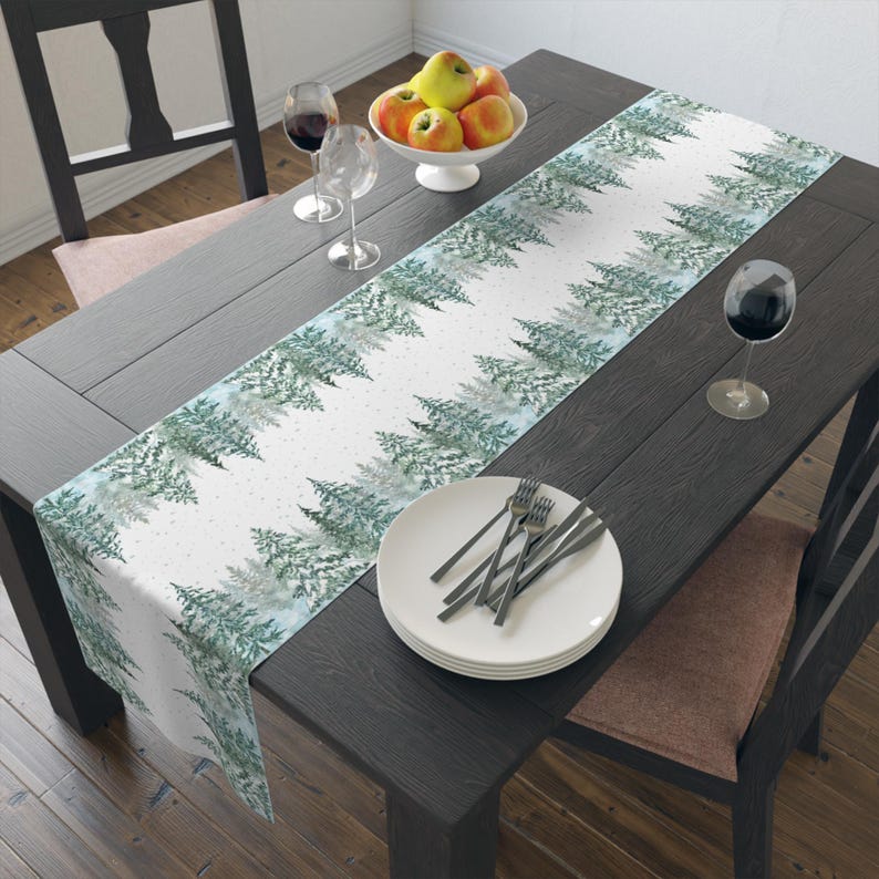 Puede incluir: Un camino de mesa blanco con un patr&oacute;n repetido de pinos verdes y azules en acuarela. El camino de mesa est&aacute; sobre una mesa de madera oscura, con platos, cubiertos y copas de vino. Un cuenco de manzanas est&aacute; en el centro.