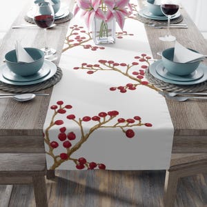 May include: White table runner with a watercolor design of red berries on brown branches. The runner is placed on a wooden table set for a meal, with teal-colored bowls, plates, and wine glasses.